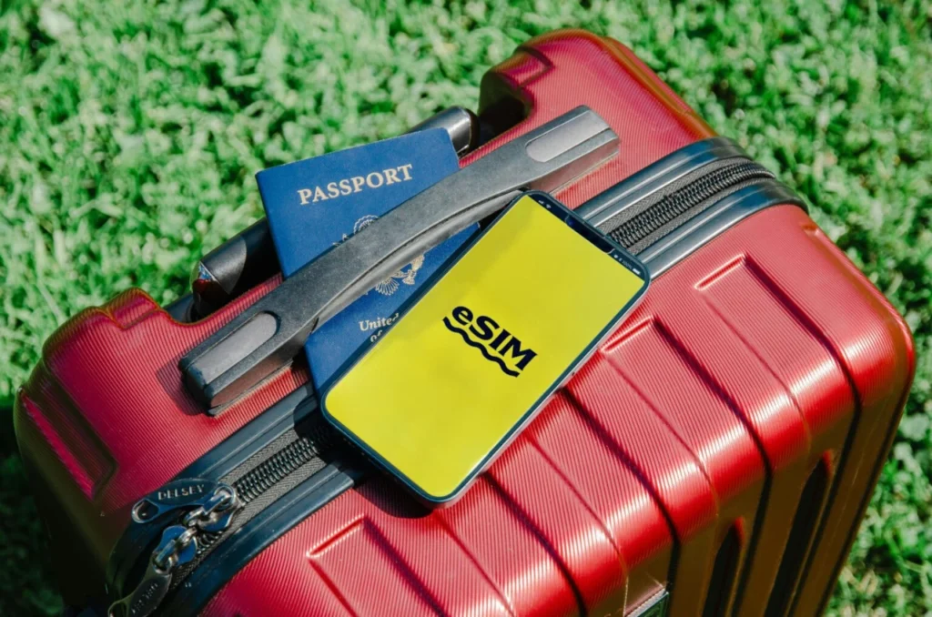 A smartphone displaying an eSIM logo resting on a red suitcase next to a blue United States passport on green grass.