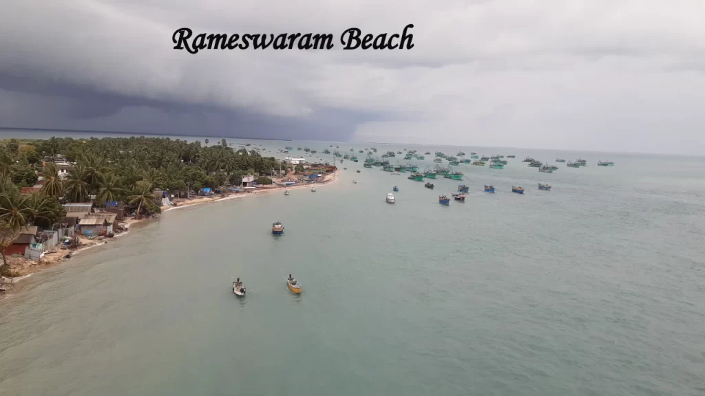 An aerial view of Rameswaram Beach captured from the Pamban Bridge, showing numerous fishing boats scattered across the turquoise ocean under a dramatic cloudy sky—a top destination to visit in TamilNadu.