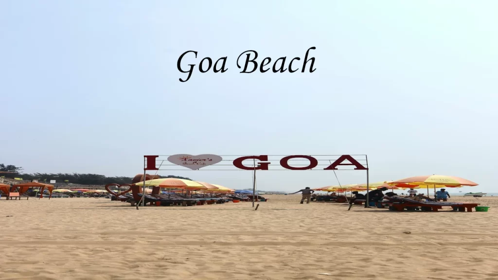 A wide sandy shore featuring sun loungers, colorful umbrellas, and a prominent red sign that reads I Love Goa.