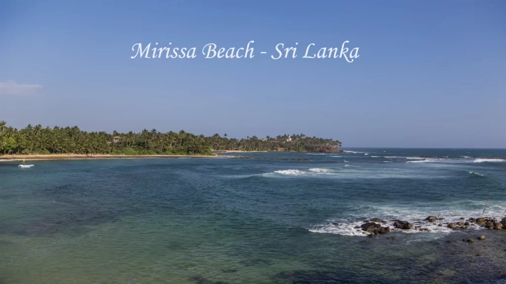 Panoramic view of Mirissa Beach in Sri Lanka showing a curved sandy shoreline, turquoise Indian Ocean waves, a fishing boat, and a lush coconut palm headland under a clear blue sky