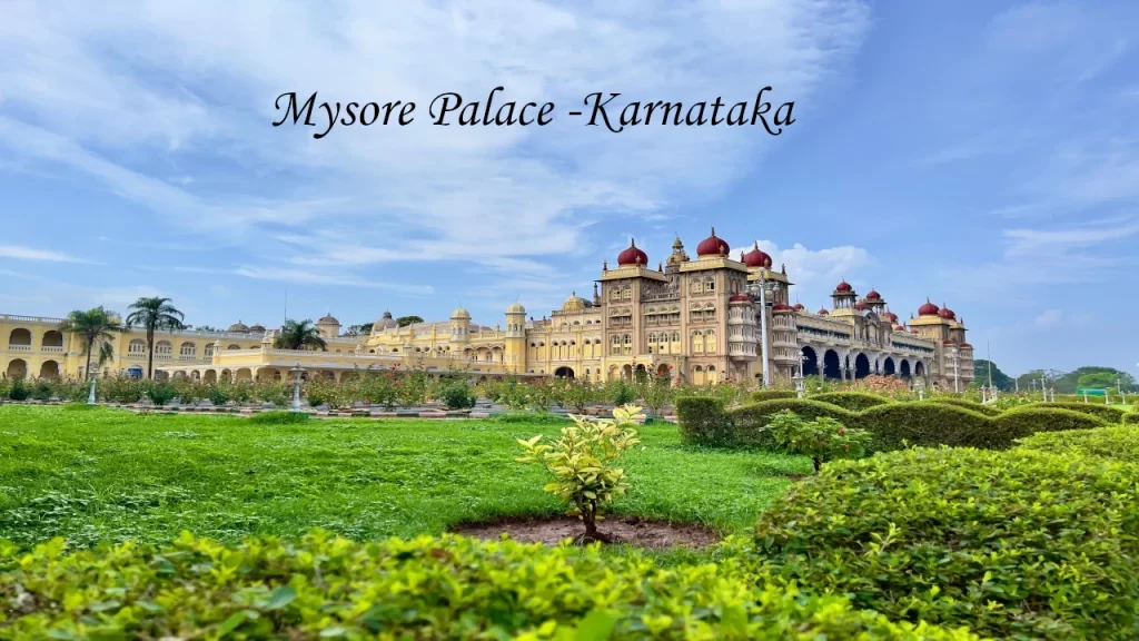 The grand exterior view of Mysore Palace in Karnataka, featuring red domes and lush green gardens under a cloudy blue sky.
