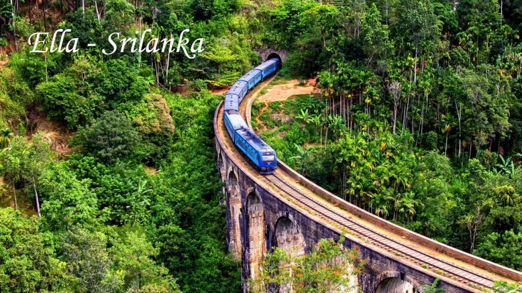 The iconic blue train passing over the historic Nine Arch Bridge in Ella along the Pekoe Trail in Sri Lanka, surrounded by dense green jungle and palm trees.