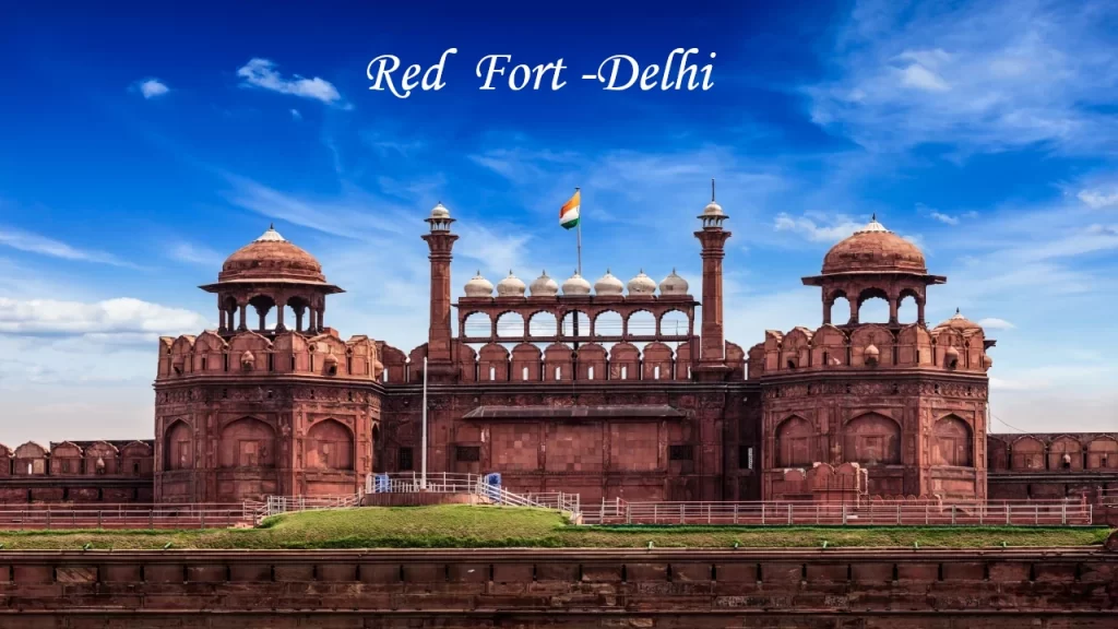 The iconic front view of the Red Fort (Lal Qila) in Delhi with the Indian national flag hoisting under a bright blue sky.