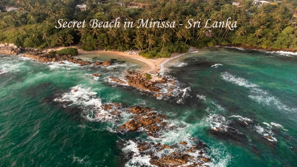 Aerial drone view of Secret Beach in Mirissa, Sri Lanka, showing turquoise Indian Ocean waters, rocky outcrops, golden sand, and lush coconut palm forest