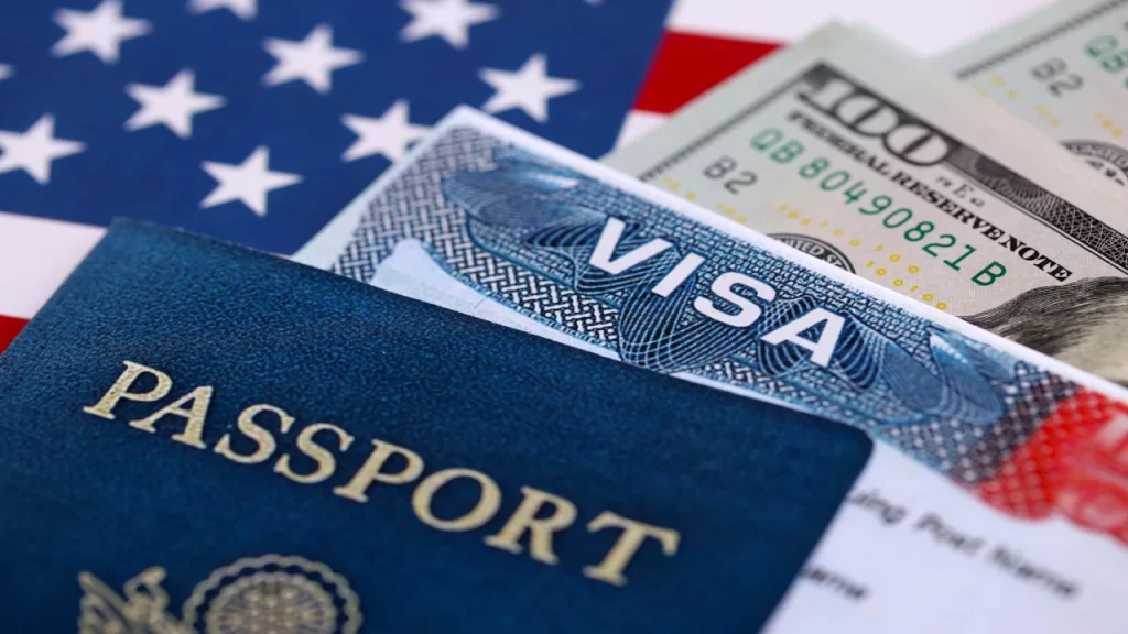 A close-up shot of an approved US tourist visa inside a passport next to one-hundred-dollar bills on an American flag background.