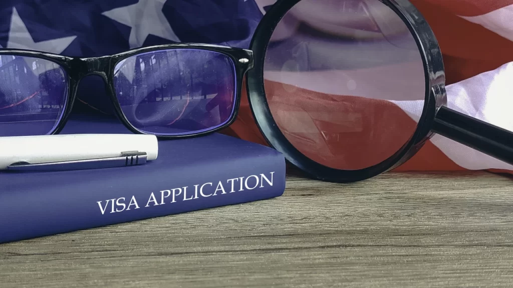 Reading glasses and a magnifying glass resting on a blue book titled Visa Application with an American flag background.
