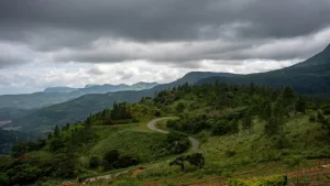 A winding asphalt road curving through lush green hills and pine trees with misty mountain ranges under dramatic storm clouds in the Knuckles region.