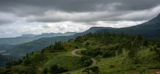A winding asphalt road curving through lush green hills and pine trees with misty mountain ranges under dramatic storm clouds in the Knuckles region.