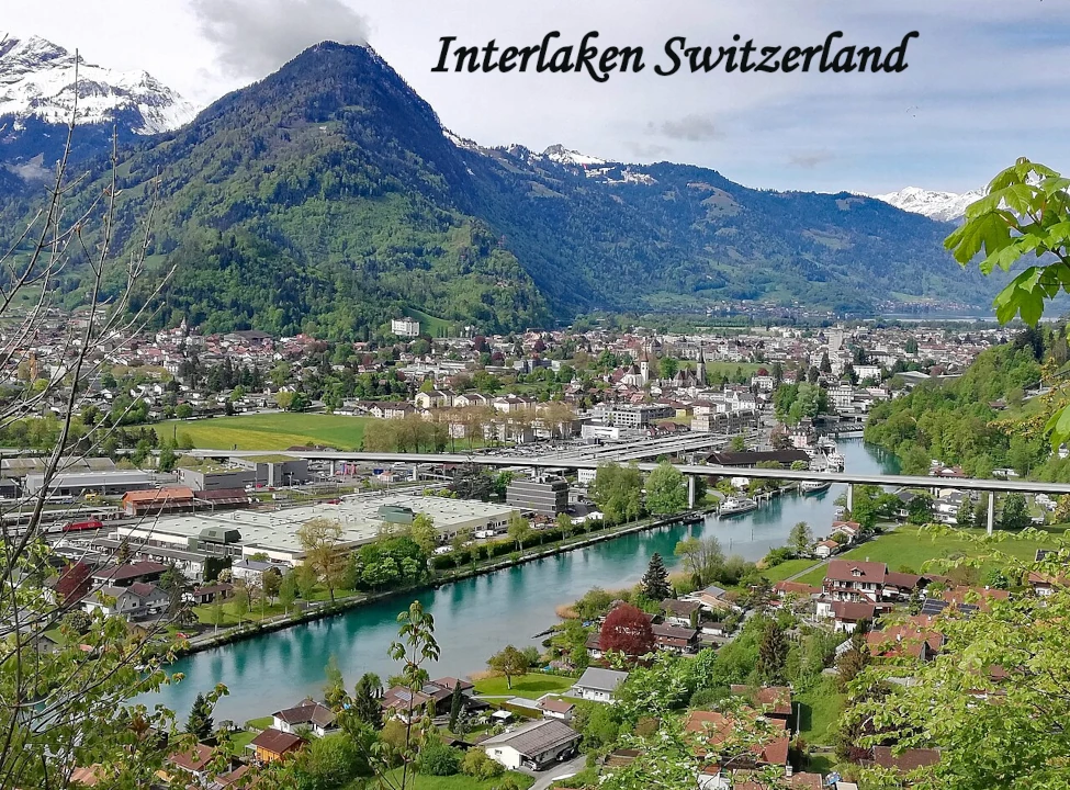 Panoramic aerial view of Interlaken town nestled between green forested mountains and snow-capped Alps, with the turquoise Aare River and a road bridge cutting through the centre of the town.