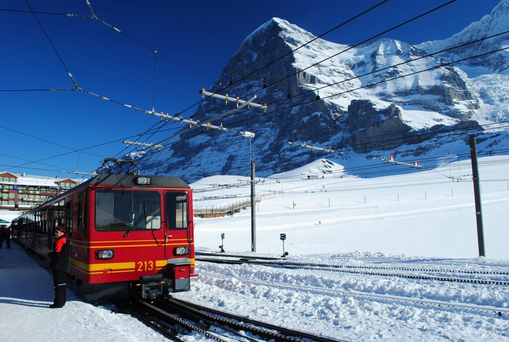 Red and yellow Jungfraubahn electric rack railway train numbered 213 stopped at a snow-covered mountain station, with the massive dark rock face of the Eiger rising steeply under a vivid blue winter sky.