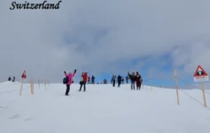 Must-Visit Tourist Places in Switzerland posing and taking photos on a vast snow-covered alpine summit in Switzerland, with red triangular warning STOP signs and rope barriers marking a safety boundary along the snowy ridge.