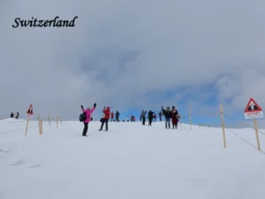 Must-Visit Tourist Places in Switzerland posing and taking photos on a vast snow-covered alpine summit in Switzerland, with red triangular warning STOP signs and rope barriers marking a safety boundary along the snowy ridge.
