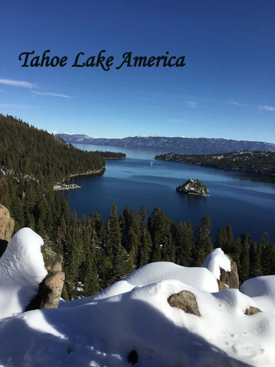 Scenic view of Lake Tahoe America with crystal-clear blue water, forested shoreline, snow-covered rocks, and surrounding mountain ranges