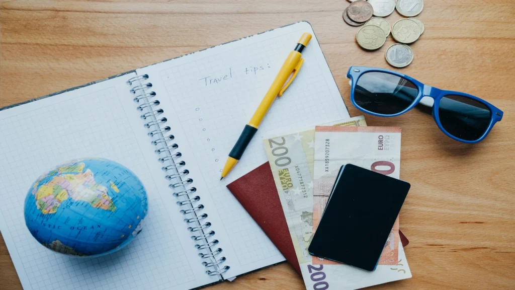 Travel checklist notebook with a globe, sunglasses, passport, Euro banknotes, and coins, representing Forex card travel planning.