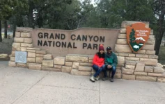 Grand Canyon National Park entrance sign with two visitors sitting beside the National Park Service sign surrounded by trees.